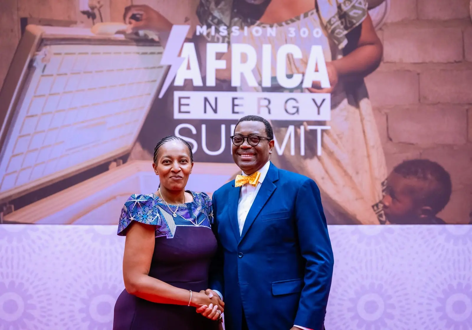 A woman shaking hands with a smiling man in a blue suit and yellow bow tie in front of an Africa Energy Summit backdrop