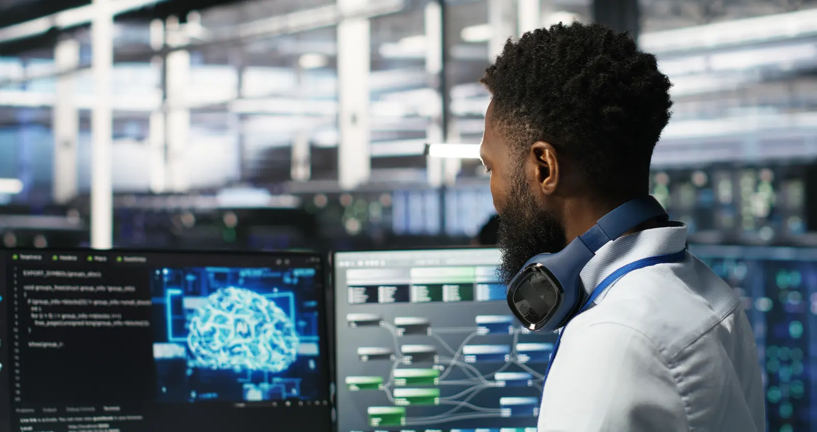 A man with headphones around his neck looks at monitors showing a glowing brain graphic and system diagrams in a data centre
