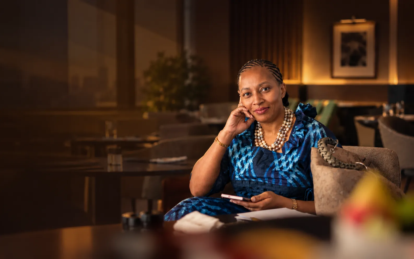 A woman seated in a warmly lit lounge, resting her chin on her hand and smiling at the camera